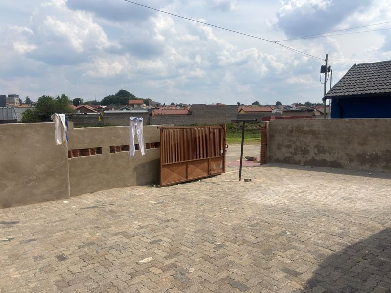 A rear exterior area featuring a paved driveway, metal gate, and unfinished boundary wall, providing access to the property.