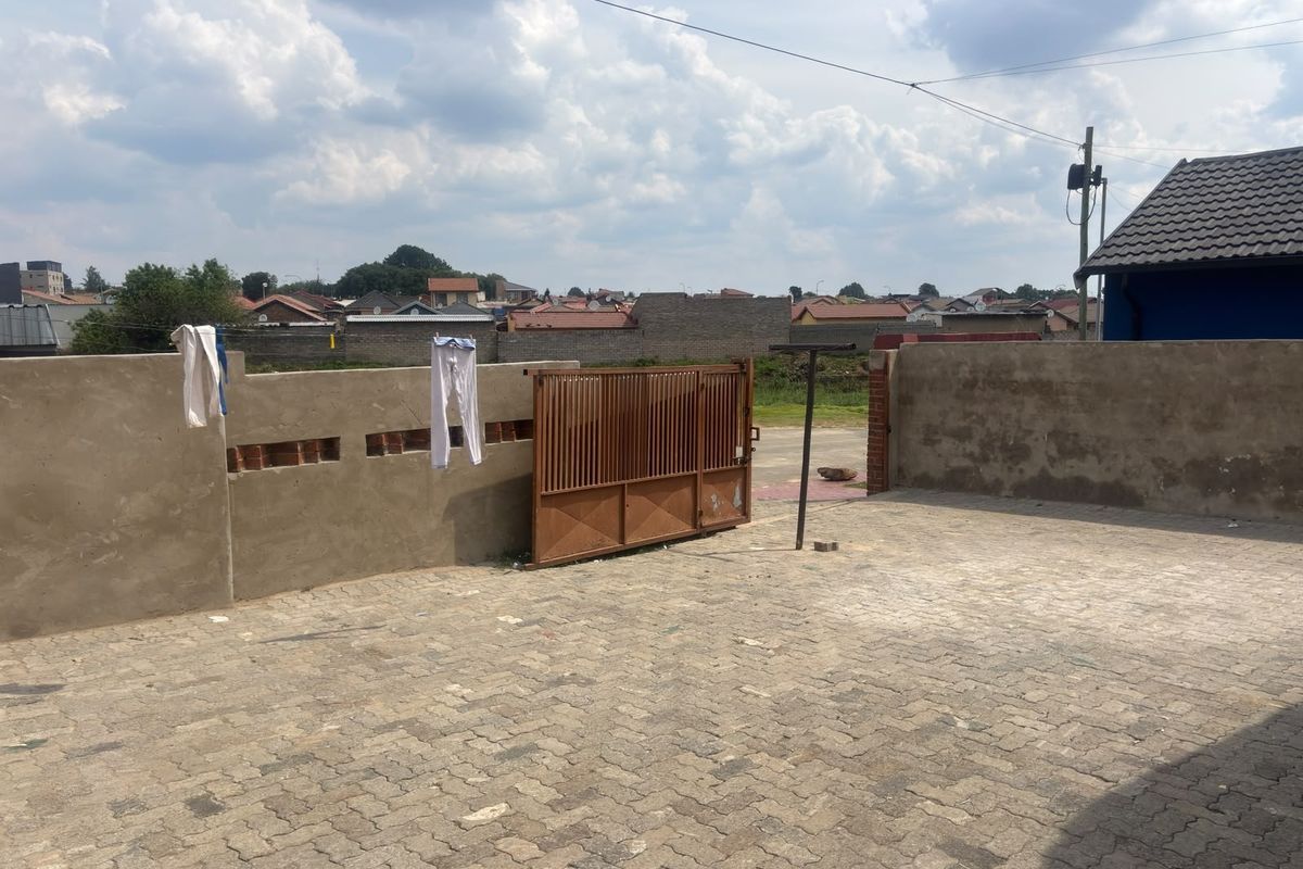 A rear exterior area featuring a paved driveway, metal gate, and unfinished boundary wall, providing access to the property.