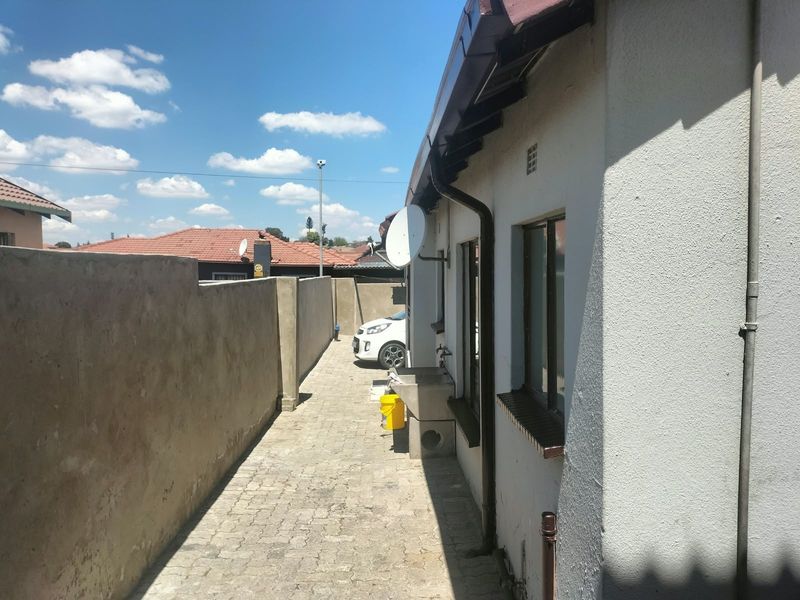 A side exterior view of the cottage featuring a paved walkway, boundary wall, and an external utility sink attached to the building.