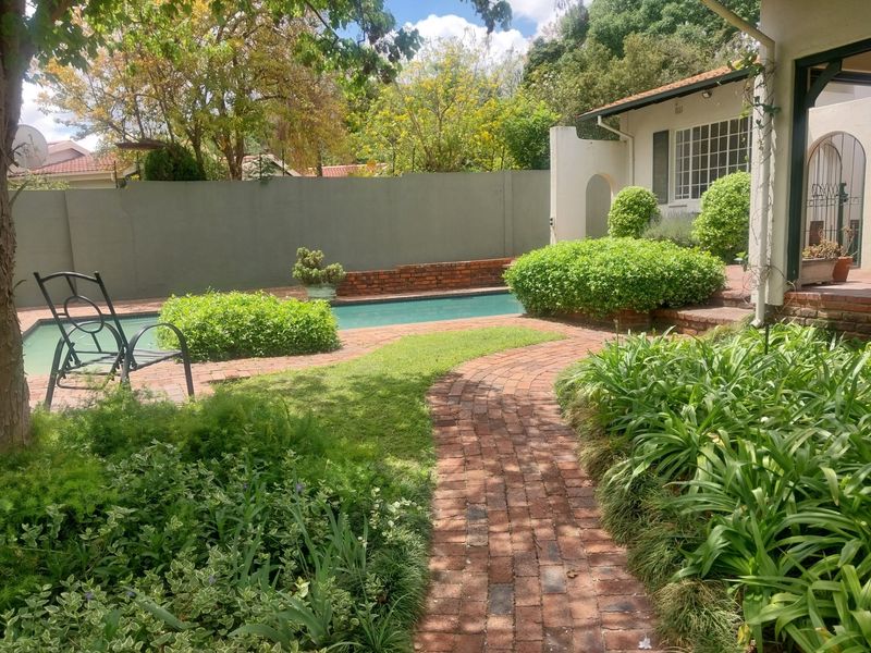 A well-maintained outdoor pool area featuring brick paving, established garden landscaping, and a secure boundary wall.