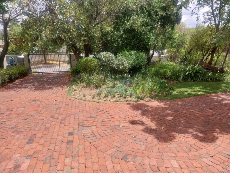 A front exterior view featuring a circular pattern brick paved driveway, mature garden landscaping, and a secure metal entrance gate.