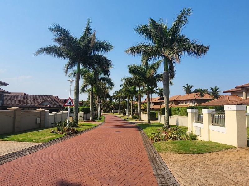 A well-maintained residential street scene featuring a paved driveway, perimeter walls, and mature palm trees within a landscaped estate setting.