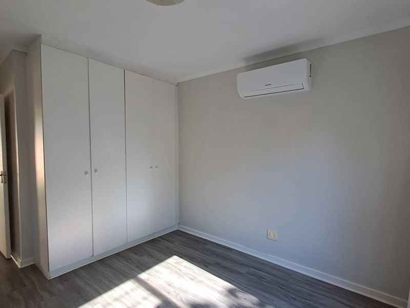A bright, neutral bedroom featuring modern laminate flooring, ample built-in cupboard storage, and a wall-mounted split-system air conditioning unit.