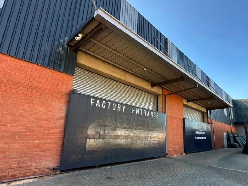 Industrial factory entrance featuring large roller shutter doors, brick and metal cladding facade, and a protective canopy over the paved loading area.
