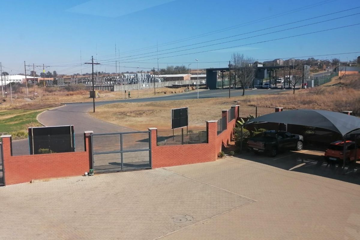 Aerial view of the industrial property entrance featuring a paved driveway, brick perimeter wall, security gate, and a shade cloth carport.