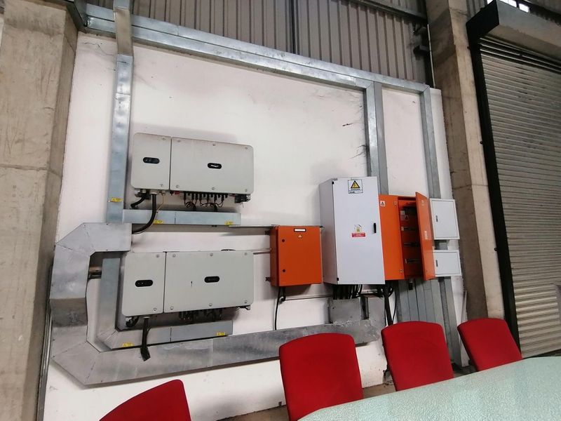 Industrial utility area featuring wall-mounted solar inverters and electrical distribution boards housed in galvanized steel trunking.