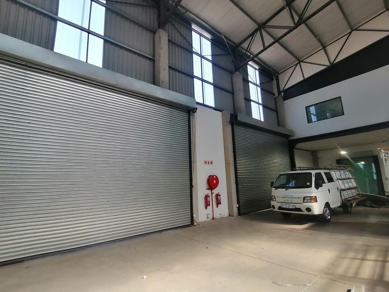 Spacious industrial warehouse unit featuring high ceilings with exposed steel trusses, large roller shutter doors, and concrete flooring.