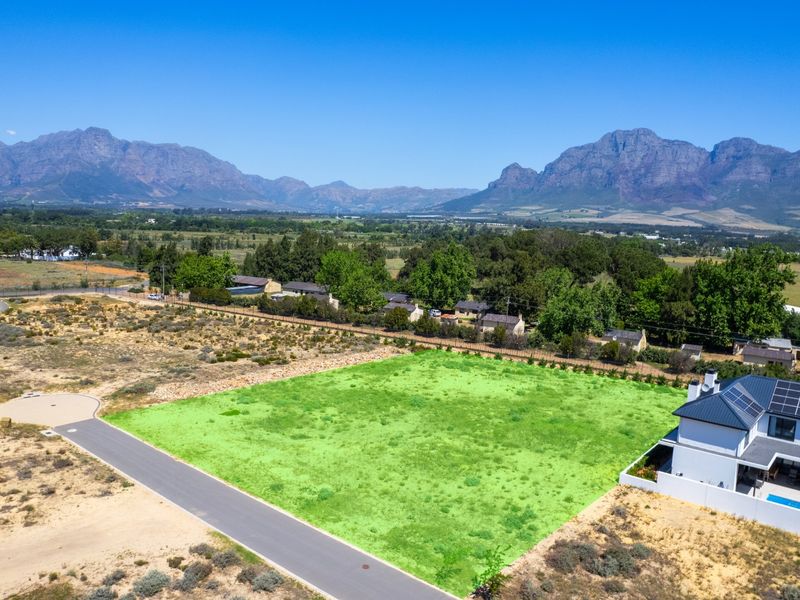 Aerial view of a vacant residential plot featuring mountain views, adjacent to established housing and paved road access.
