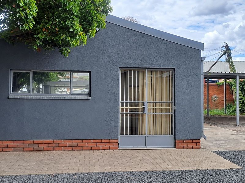 A well-maintained office exterior featuring a dark grey rendered facade, brick base, paved entrance area, and an adjacent covered carport.