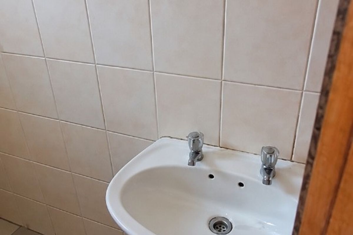 A functional office bathroom featuring a wall-mounted ceramic basin, dual-handle taps, neutral wall and floor tiling, and a simple wall-mounted mirror.