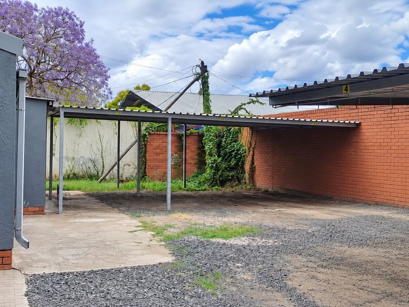 Outdoor parking area featuring a covered carport structure with brick boundary walls and a combination of paved and gravel surfaces.