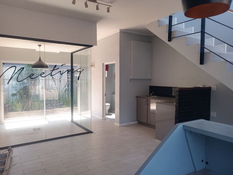 Modern open-plan commercial space featuring a glass-enclosed meeting room, kitchenette with stainless steel cabinetry, and a staircase leading to the upper level.