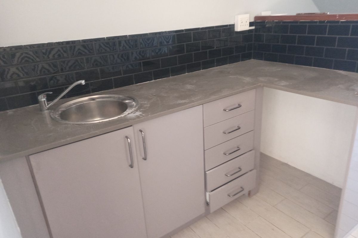 A compact kitchen area featuring grey built-in cabinetry, a stainless steel sink, and a dark subway tile backsplash with stone countertops.