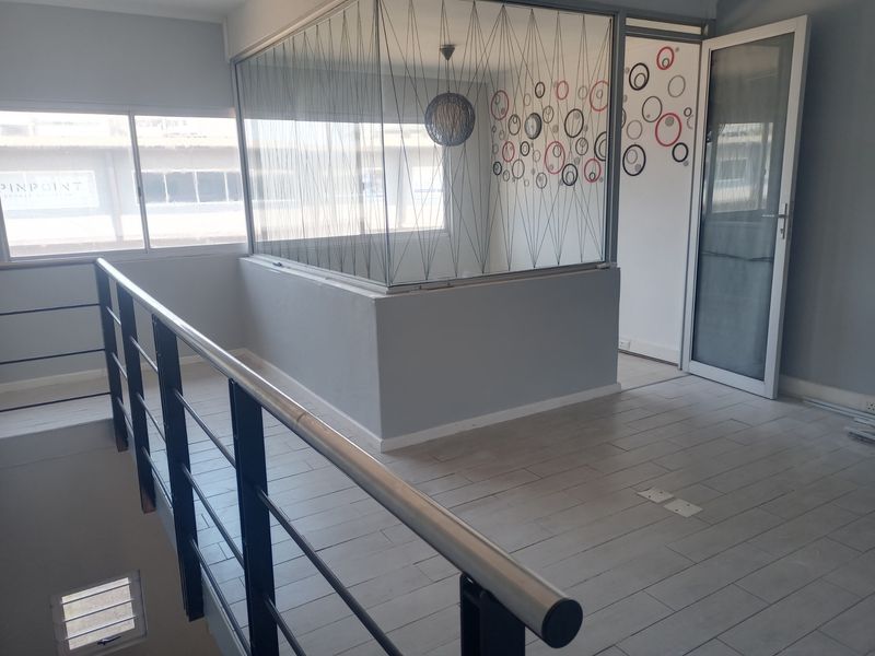 An open-plan office space featuring a glass-partitioned meeting room, tiled flooring, and a metal staircase railing, suitable for a professional commercial environment.