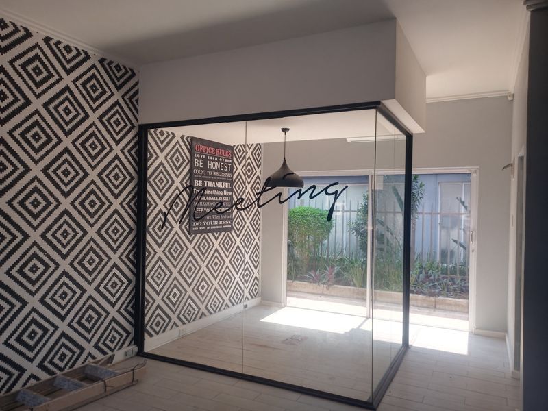 A modern, glass-enclosed office meeting room featuring black metal framing, patterned wallpaper, and tiled flooring, offering a bright and professional workspace.