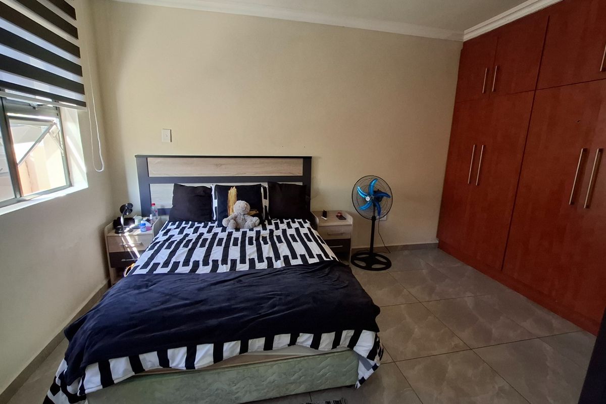 A functional bedroom featuring tiled flooring, built-in wooden cupboards, and a window fitted with modern roller blinds.