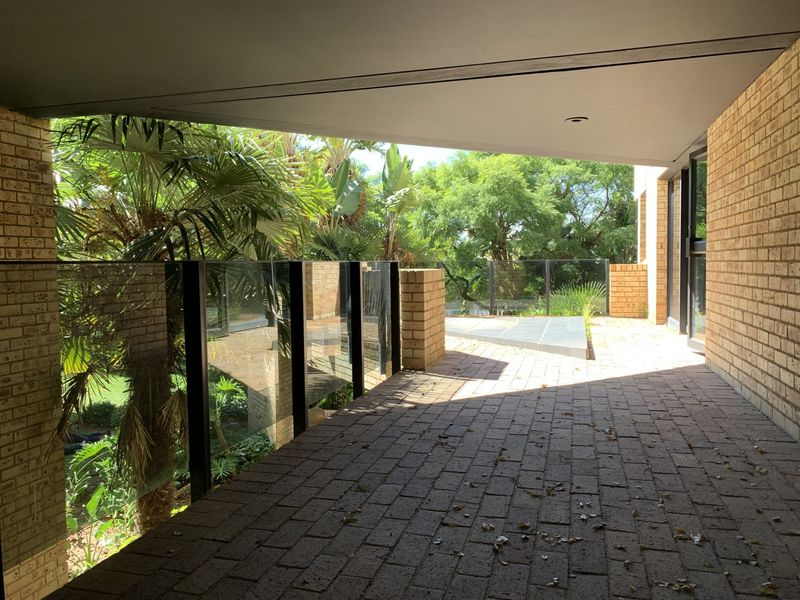 A spacious covered patio featuring brick paving, a modern glass balustrade, and views of the surrounding garden.