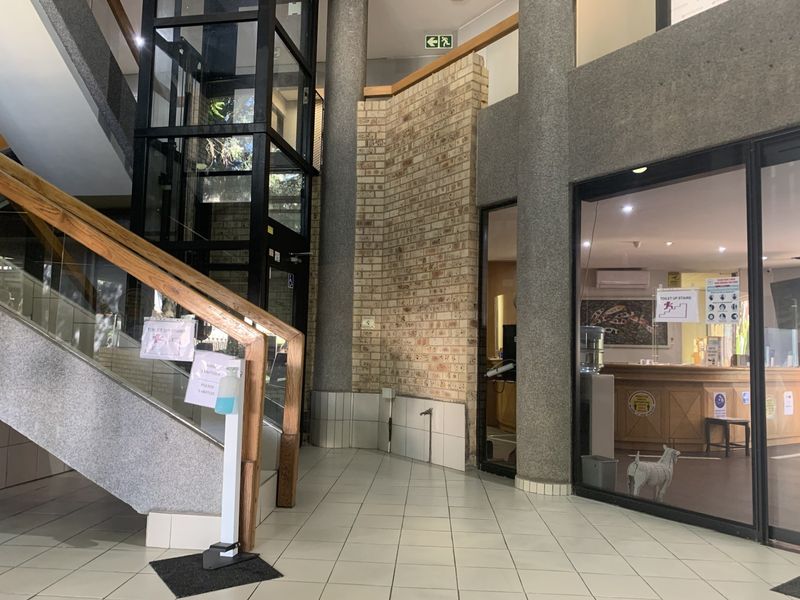 Commercial building entrance lobby featuring a glass elevator, concrete columns, exposed brickwork, and a staircase with a wooden handrail.