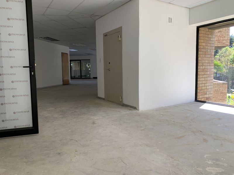 An empty, open-plan commercial office space featuring a suspended ceiling, exposed concrete flooring, and a large window with brick detailing.