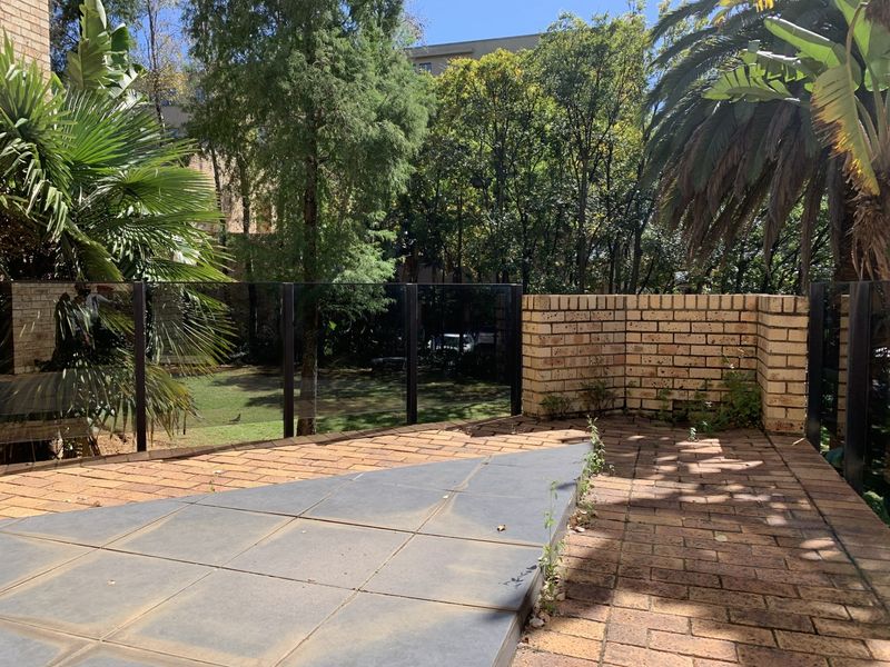 An outdoor patio area featuring brick paving, a glass balustrade, and a brick retaining wall, overlooking a landscaped garden space.