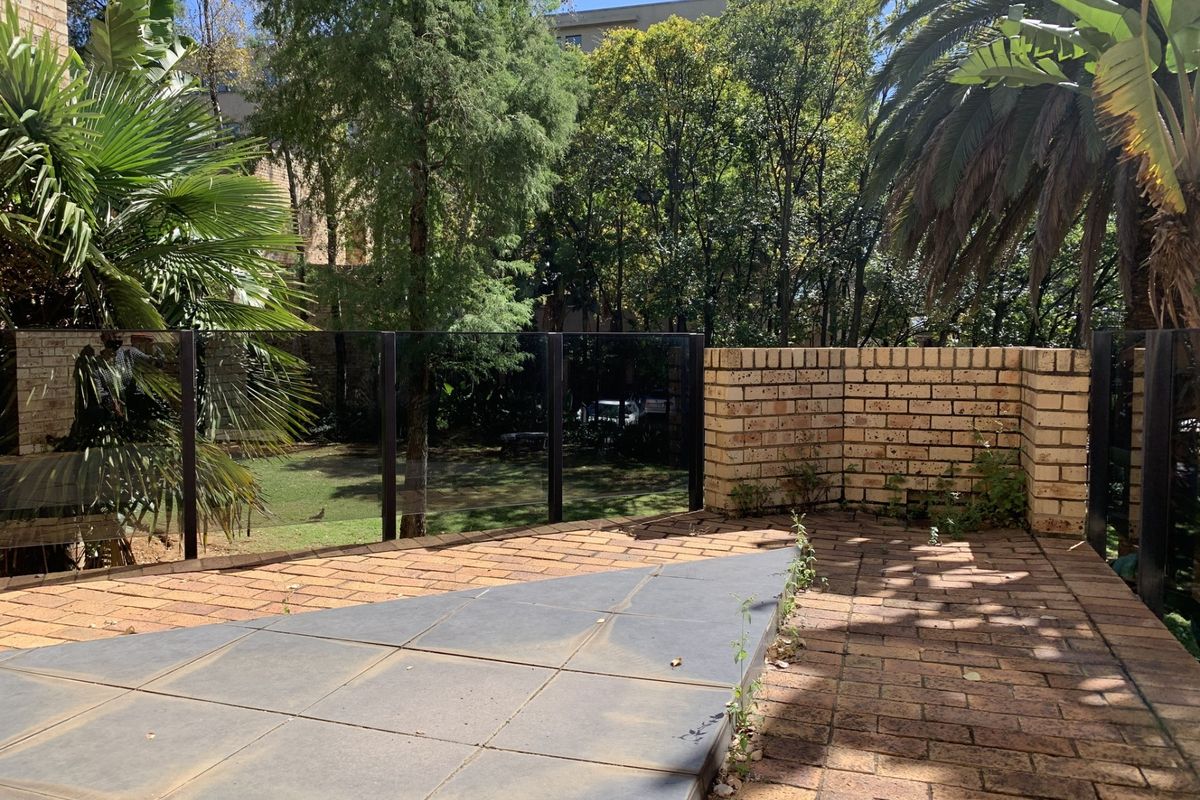 An outdoor patio area featuring brick paving, a glass balustrade, and a brick retaining wall, overlooking a landscaped garden space.