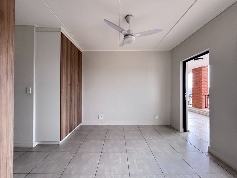 A bright, empty bedroom featuring modern tiled flooring, built-in wooden cupboards, a ceiling fan, and direct access to an outdoor balcony.