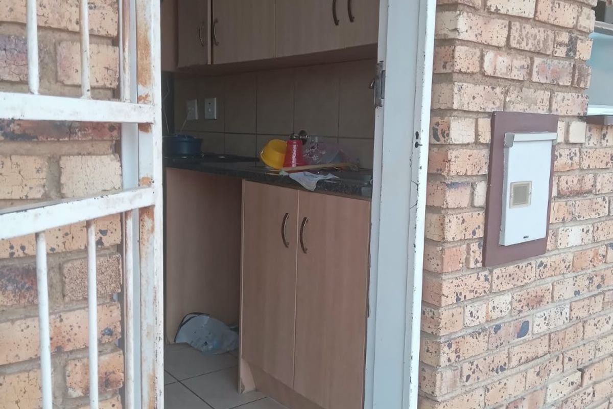 A compact kitchen area featuring laminate cabinetry and tiled flooring, accessed via an exterior security gate in a brick-walled apartment setting.