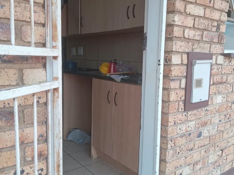 A compact kitchen area featuring laminate cabinetry and tiled flooring, accessed via an exterior security gate in a brick-walled apartment setting.