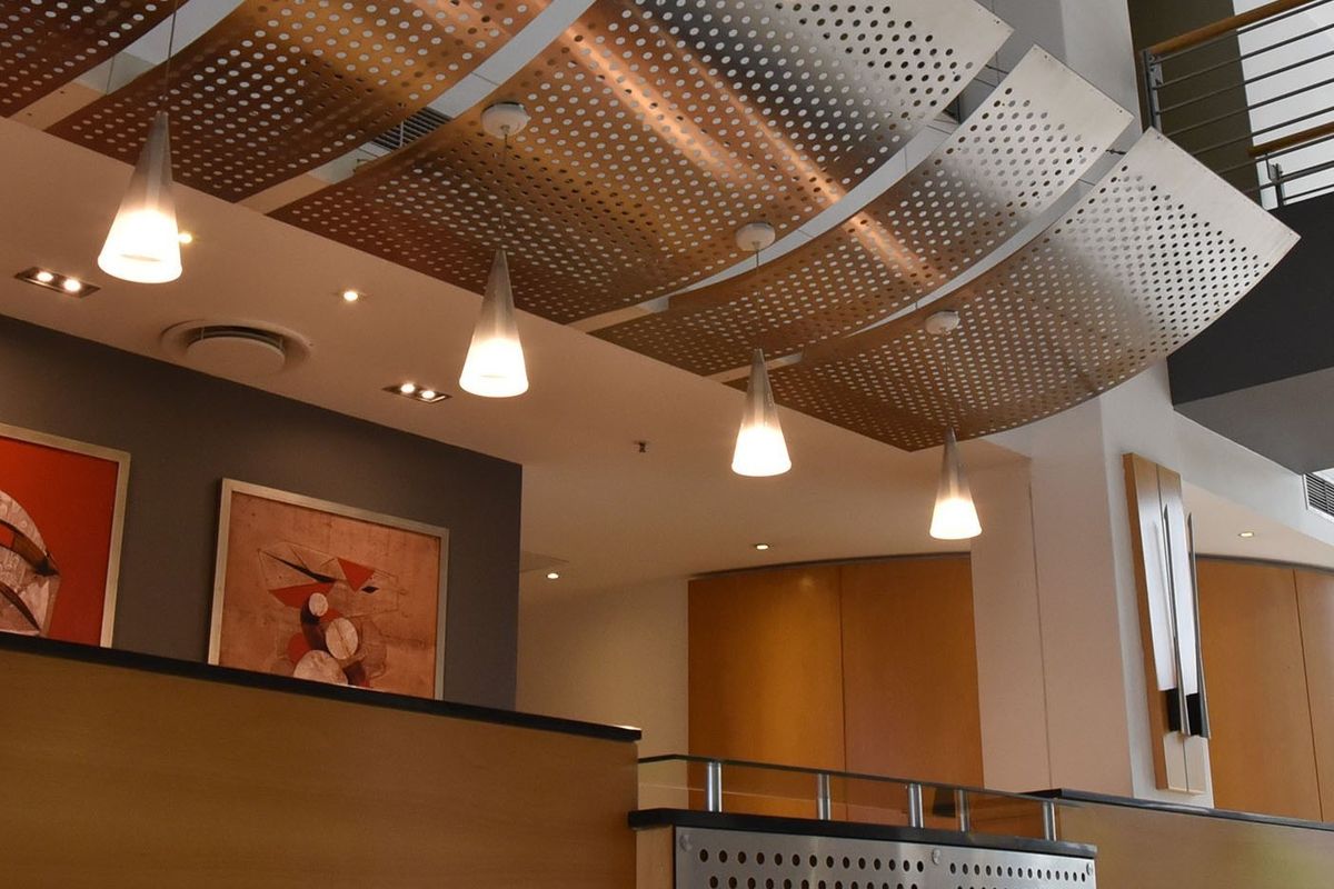 Modern commercial lobby entrance featuring architectural perforated metal ceiling panels, warm timber veneer wall finishes, and contemporary pendant lighting.