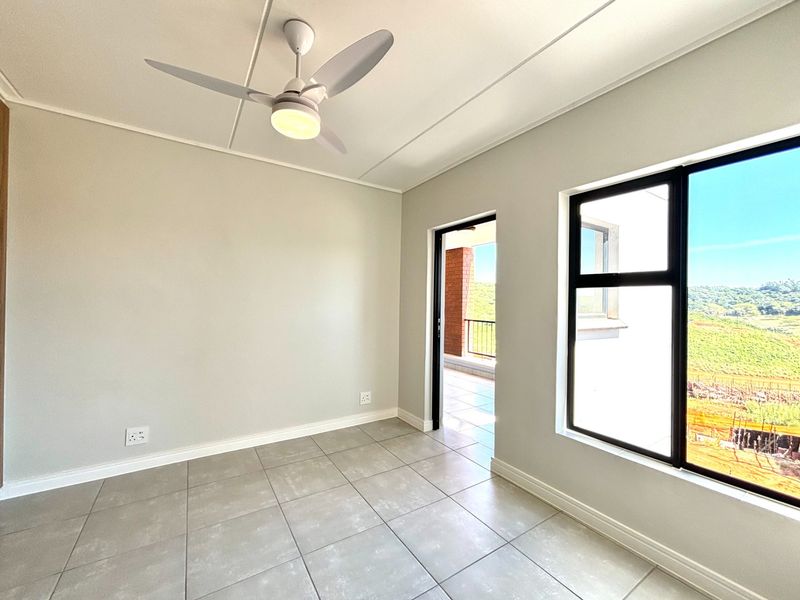 A bright, empty bedroom featuring neutral tiled flooring, a modern ceiling fan, and large windows with access to an adjoining balcony.