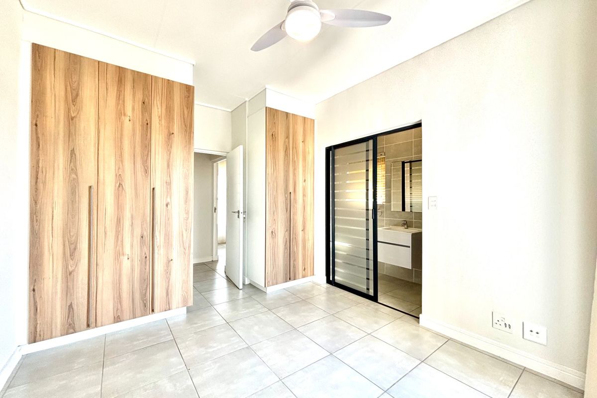A bright, empty bedroom featuring modern wood-finish built-in cupboards, tiled flooring, a ceiling fan, and direct access to an en-suite bathroom.
