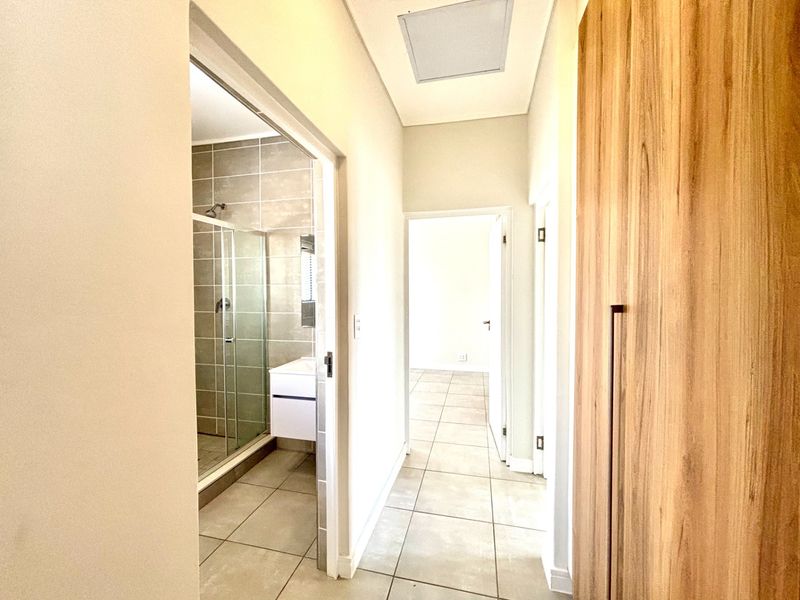 A bright, tiled hallway featuring a built-in wooden cupboard and direct access to an adjacent bathroom with a modern shower and vanity.