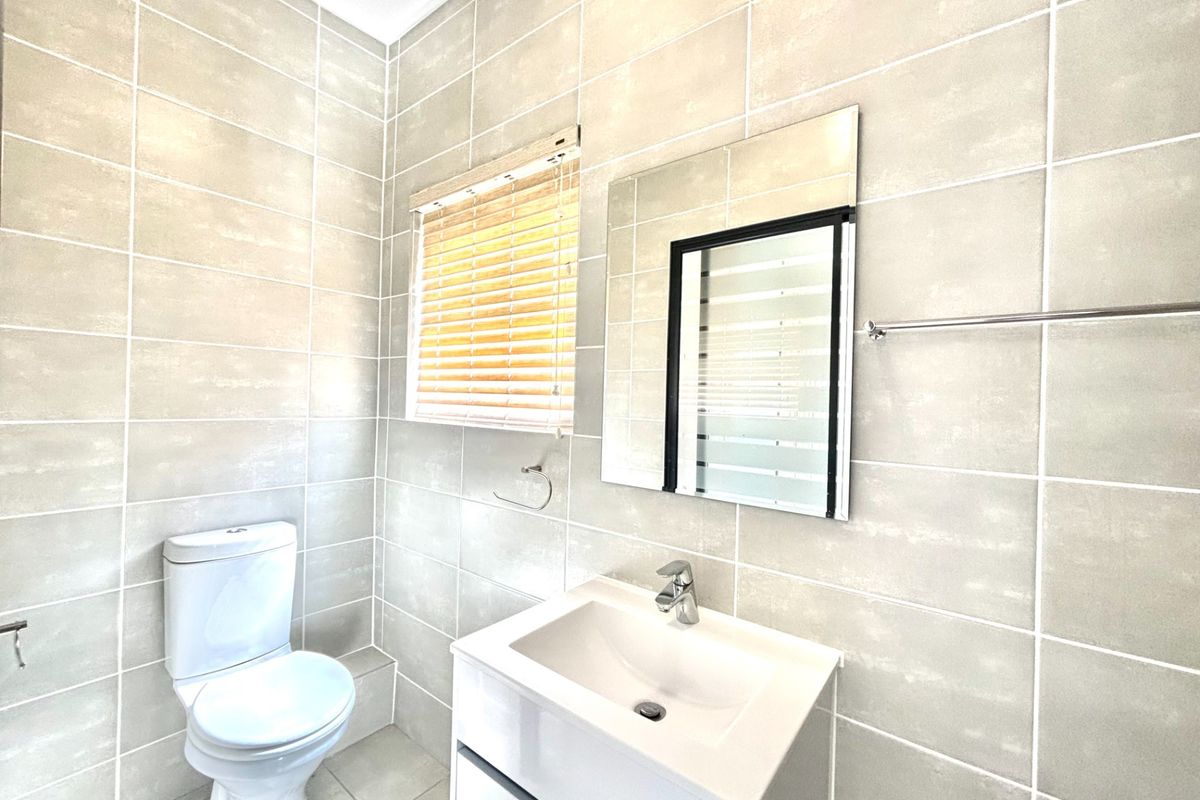 A clean, modern bathroom featuring neutral wall tiles, a white vanity with basin, a wall-mounted mirror, and a toilet.