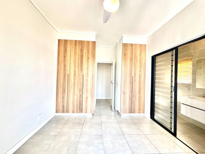 A bright, empty bedroom featuring modern wood-finish built-in cupboards, tiled flooring, a ceiling fan, and direct access to an en-suite bathroom.