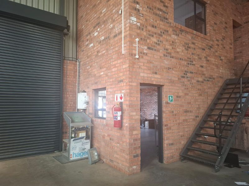 Industrial utility space featuring exposed brick walls, a metal staircase, roller shutter door, and essential safety infrastructure including a fire extinguisher and distribution board.