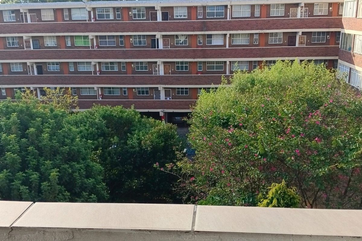 View of the rear brick facade of a multi-story apartment complex, featuring established trees and a balcony foreground.