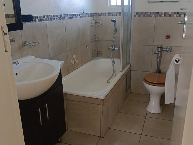 A functional bathroom featuring a vanity cabinet, bathtub with glass screen, and a toilet, finished with neutral floor and wall tiling.