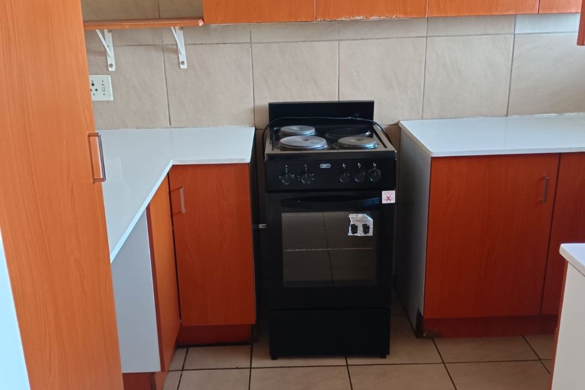 A functional kitchen featuring wood-look cabinetry, white laminate countertops, a tiled backsplash, and a freestanding stove with ceramic tile flooring.