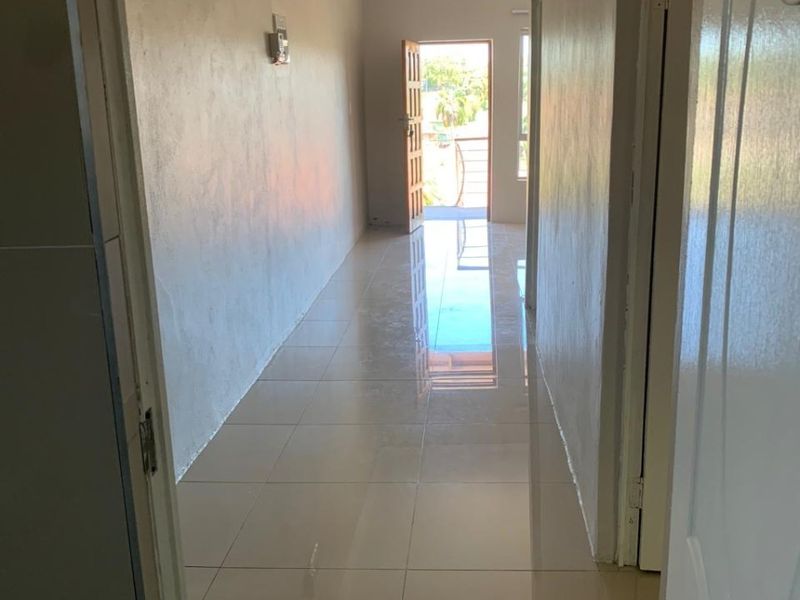 A clean, empty hallway featuring neutral tiled flooring and simple ceiling lighting, leading towards an open exterior doorway.