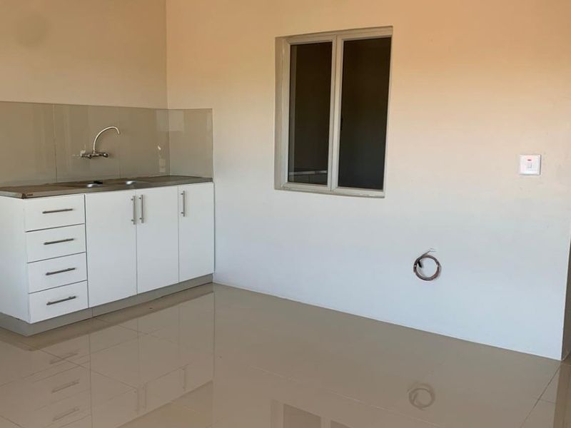 A clean, minimalist kitchen area featuring white built-in cabinetry, a stainless steel sink, tiled flooring, and neutral wall finishes in a bright, open space.