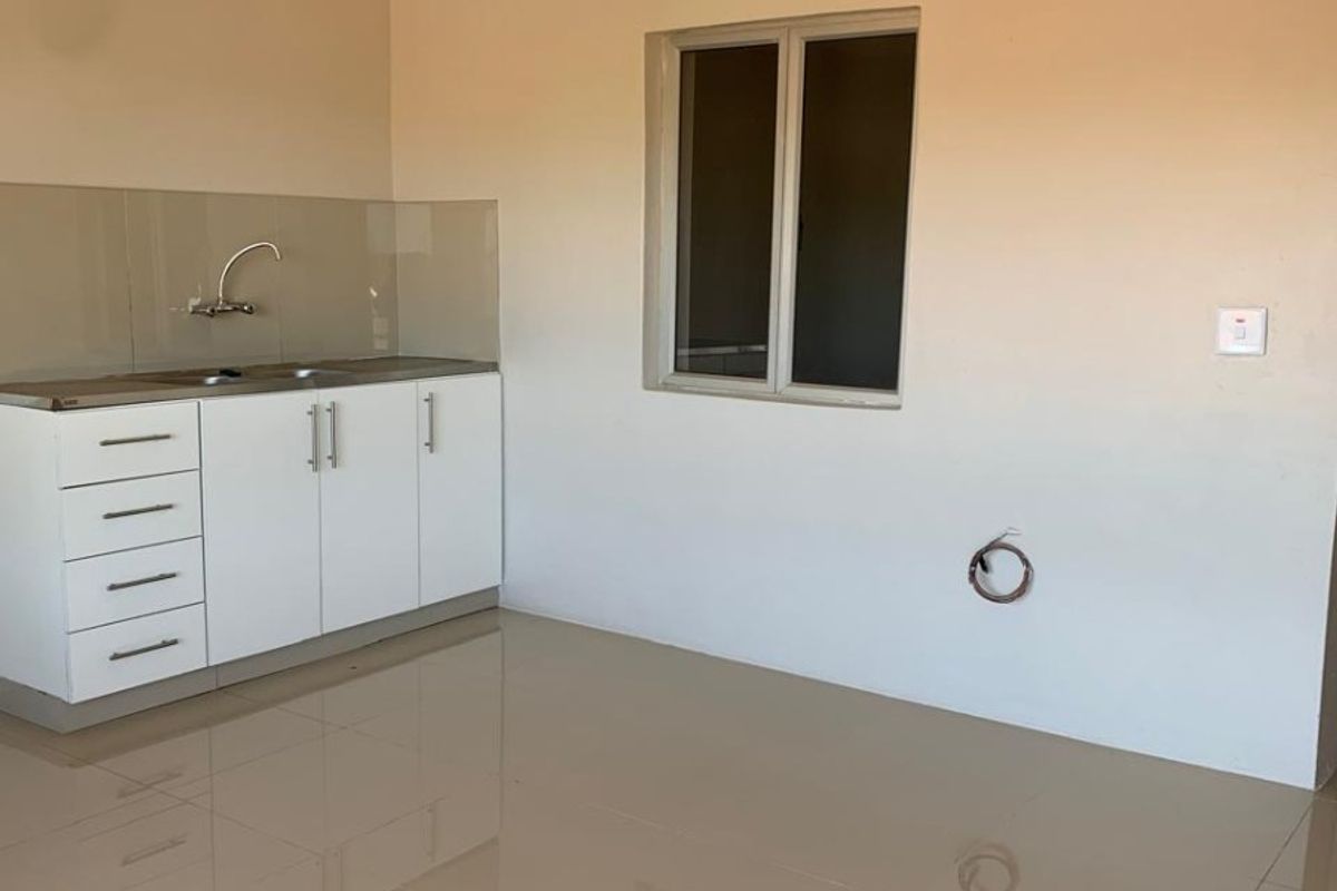 A clean, minimalist kitchen area featuring white built-in cabinetry, a stainless steel sink, tiled flooring, and neutral wall finishes in a bright, open space.