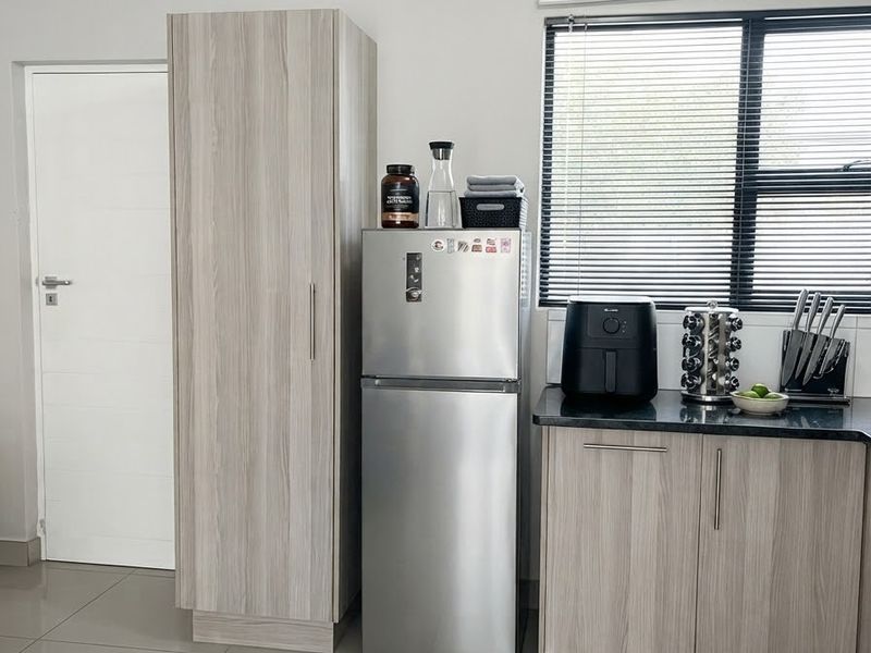 A modern kitchen area featuring light wood-grain cabinetry, dark stone countertops, large format floor tiles, and a window with fitted venetian blinds.