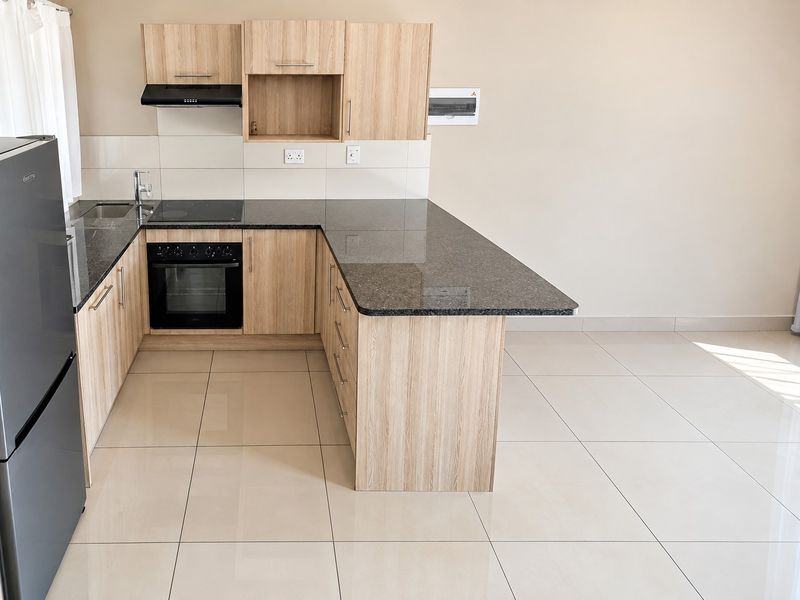 Modern kitchen featuring light wood-look cabinetry, dark granite countertops, integrated electric hob and oven, and neutral tiled flooring in an open-plan layout.