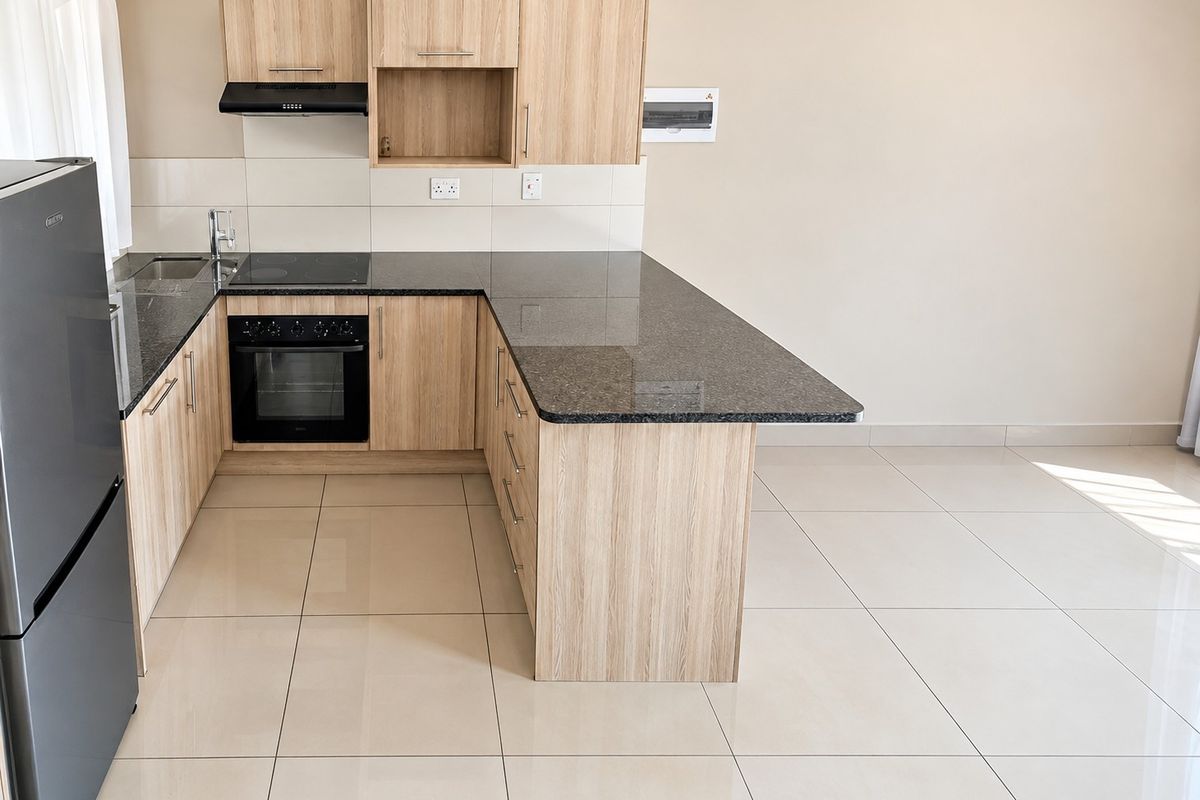 Modern kitchen featuring light wood-look cabinetry, dark granite countertops, integrated electric hob and oven, and neutral tiled flooring in an open-plan layout.