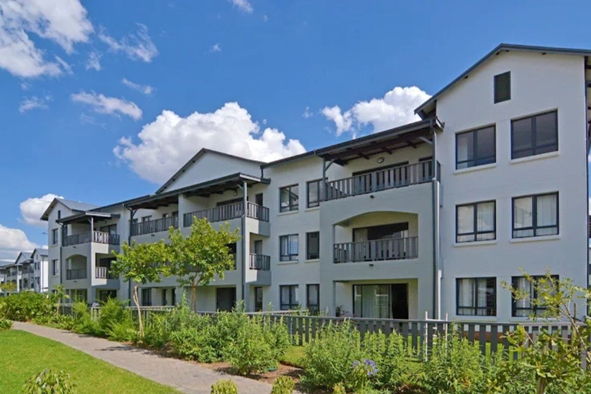A well-maintained apartment complex featuring multiple balconies, a paved walkway, and landscaped garden areas under a clear blue sky.
