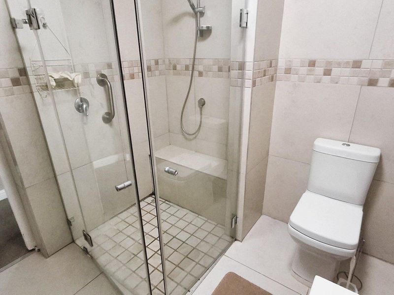 A functional bathroom featuring a glass-enclosed shower with a built-in seat, grab rails, and neutral ceramic tiling throughout.