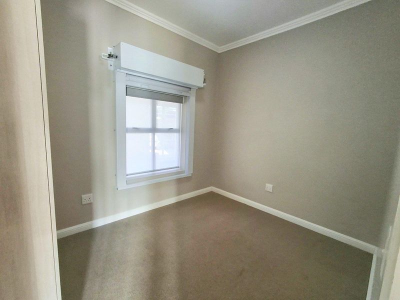 A clean, empty bedroom featuring neutral wall tones, carpeted flooring, and a window equipped with a functional roller shutter.