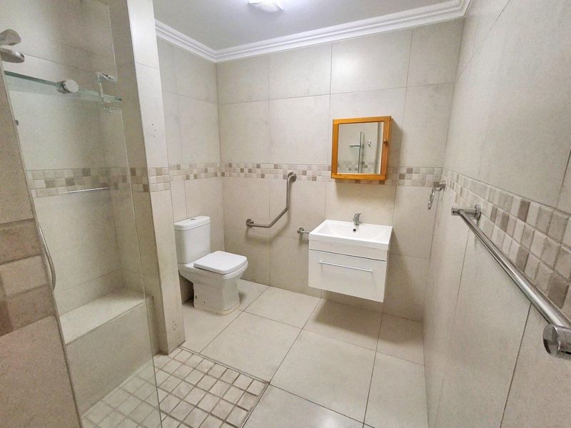 A clean, modern bathroom featuring a walk-in shower with glass screen, wall-mounted vanity, and accessible grab rail, finished with neutral floor-to-ceiling tiling.