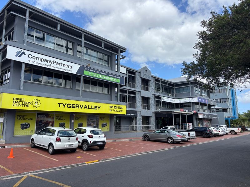 A multi-story commercial office building featuring a paved parking area, prominent storefront signage, and modern grey facade finishes.