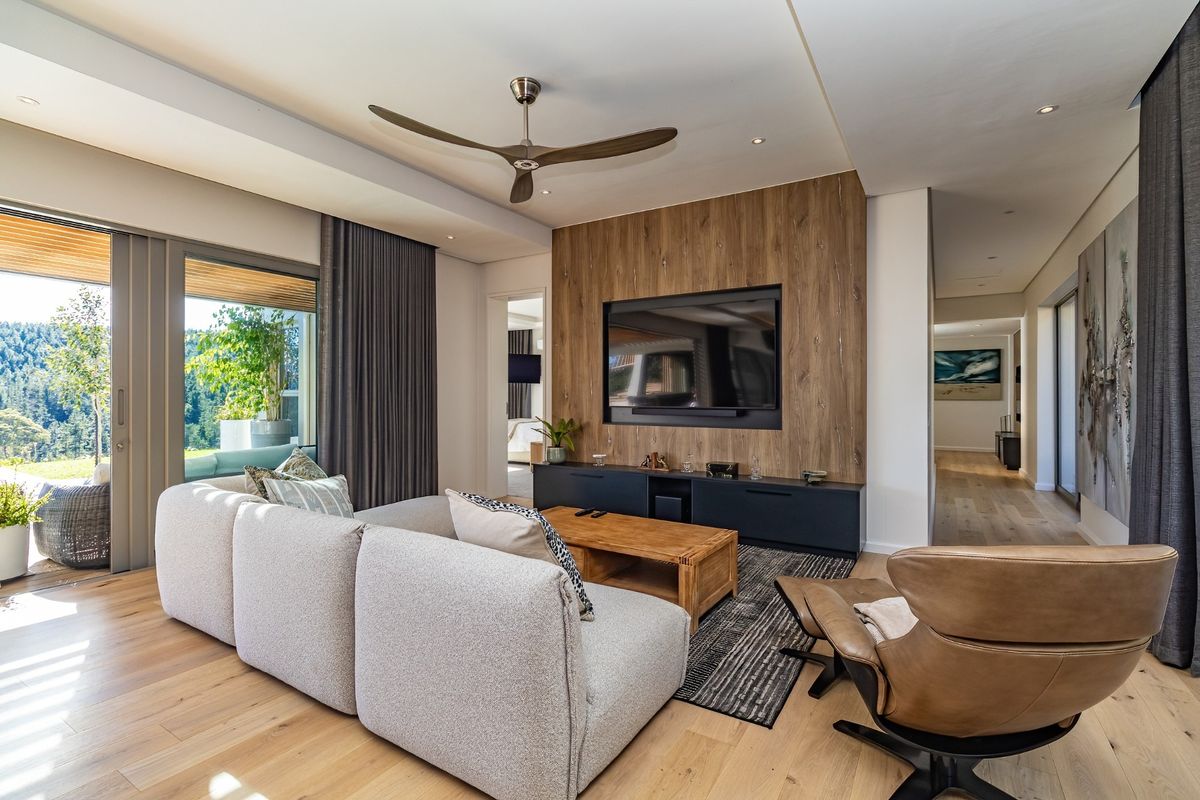 Modern living room featuring hardwood flooring, a timber feature wall with integrated media console, ceiling fan, and large sliding doors opening to a scenic view.